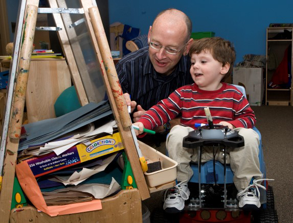 a boy on the baby robot and a man draw on an easel