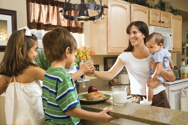 Mom holding a baby and serving food to kids in the kitchen