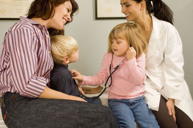 Mamá con su niño pequeño y su niña pequeña en la consulta con una doctora mientras la niña prueba el estetoscopio en el niño