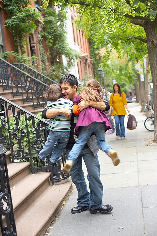 Papá abraza a sus niño y a su niña llegando de la guardería o jardín de infancia en la entrada de su casa mientras la mamá les observa cargando sus bultos/morrales