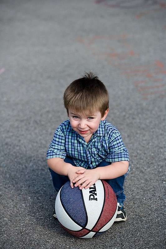 Niño pequeño sonriente agachado sobre sus piés abrazando una pelota de baloncesto en el suelo