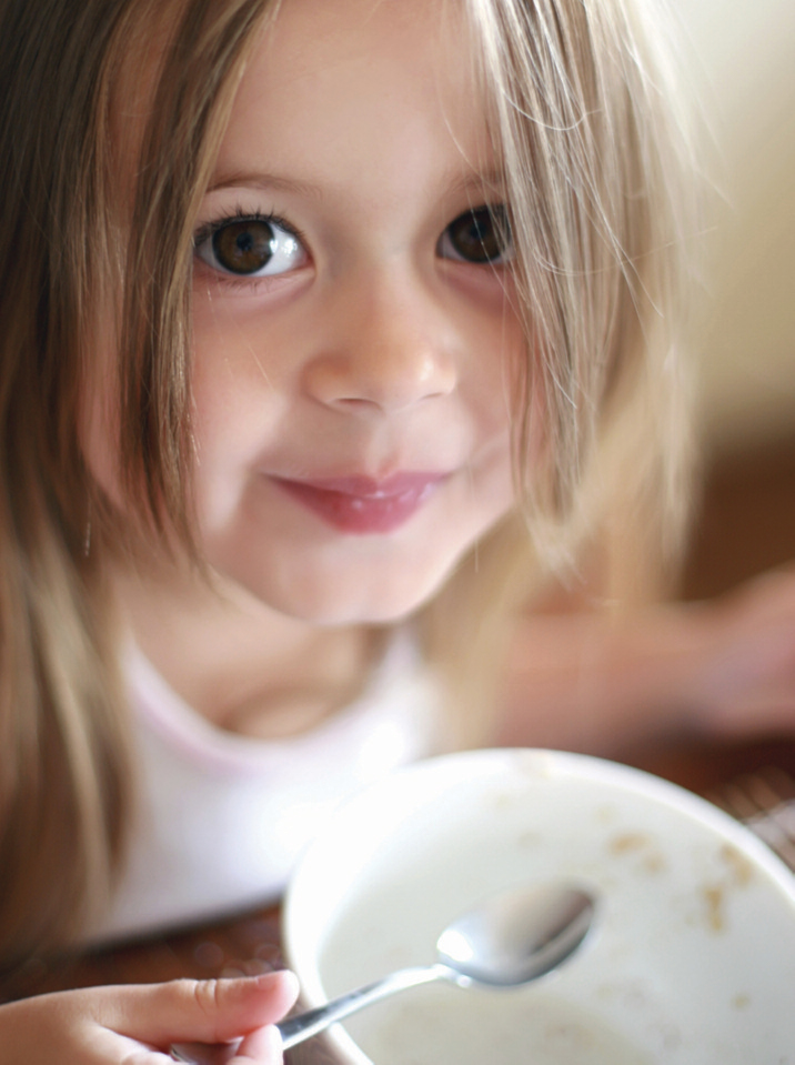 Niña pequeña sonriente sostiene una cuchara sobre un bol con restos de cereal y leche