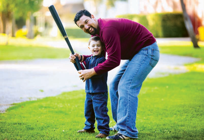 Papá e hijo sonrientes al bate con ambas manos del papá sosteniendo las de su hijo