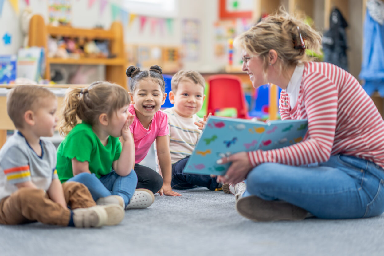 Grupo de niñas y niños pequeños/pequeñas sonríen mientran leen un libro sentados en el suelo frente a su maestra en una guardería