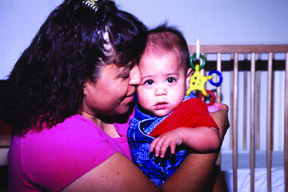 Mamá carga a su bebé quien presta atención mientras su mama agita un sonajero en su mano