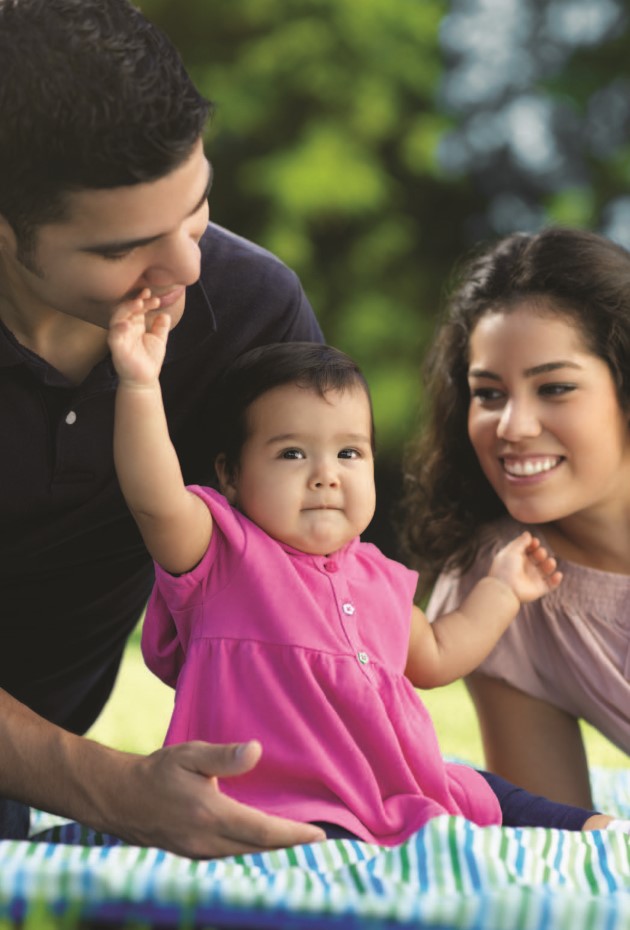 Bebé sentada en manta en un jardín o parque, alzando sus manos, tocando la boca de su papá, con papá sosteniédola por detrás y mamá observando