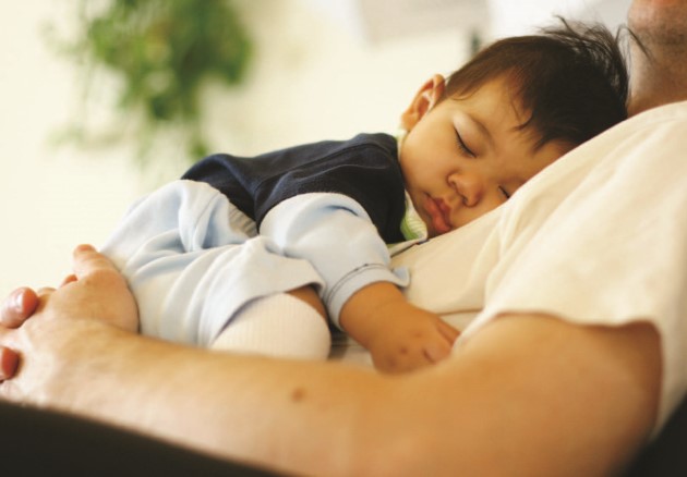 Bebé durmiendo, con brazo extendido, acostado sobre el torso de un adulto recostado en una silla o sofá