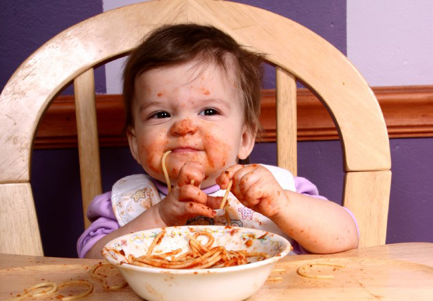 Niña pequeña usa sus manos para comer pasta con salsa de tomate sentada en su silla de comer