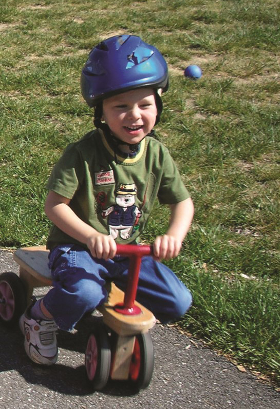 Niño pequeño con casco azul sentado en un patinete (scooter) de cuatro ruedas con las manos en el manubrio