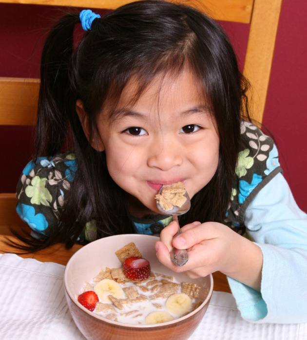 Niña pequeña come de un bol con cereal, leche y frutas utlizando una cuchara