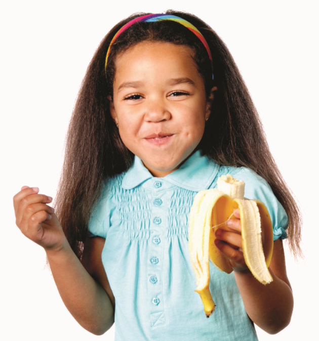 Niña pequeña sonríe y mastica un trozo de banana/cambur al que sostiene con su mano