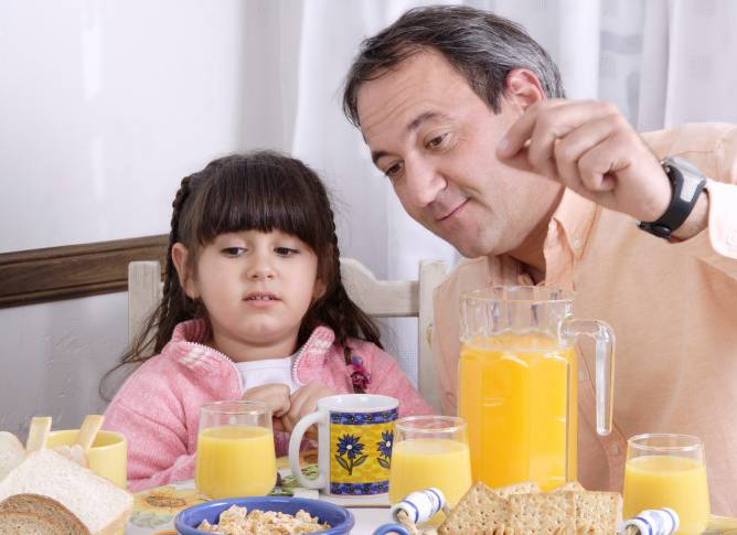 Niña pequeña con expresión un poco seria sentada en la mesa frente a un jugo/zumo de naranja con su papá