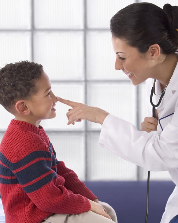 Niño pequeño sonríe en la consulta con una doctora quien le toca la punta de la nariz