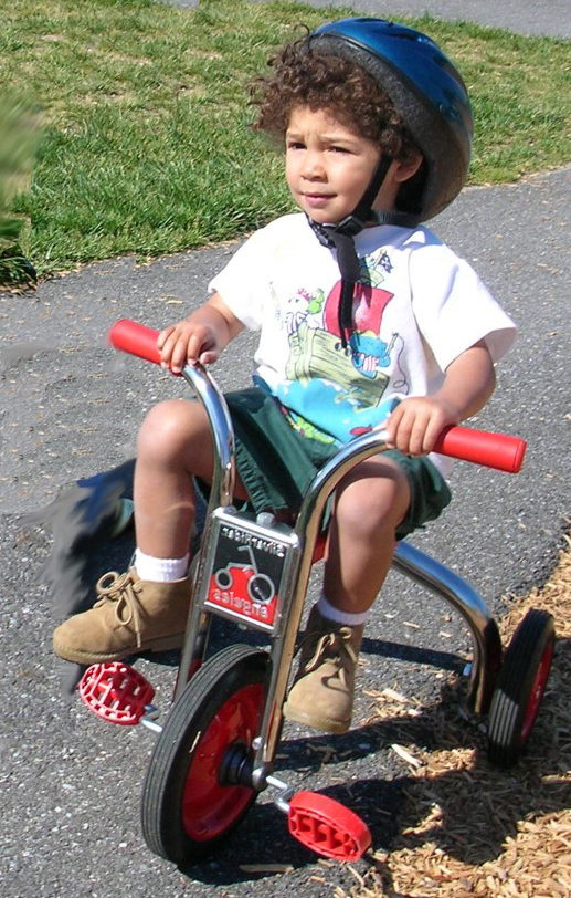 Niño pequeño con casco azul pedalea sobre su triciclo con pedales y asideras de color rojo