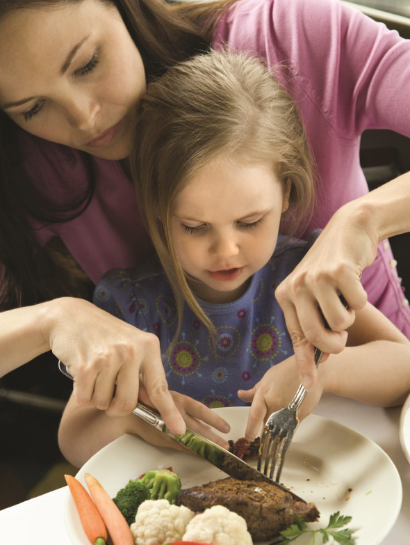 Mamá e hija pequeña cortan juntas la comida usando tenedor y cuchillo