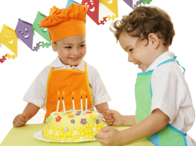 Dos niños pequeños sonrientes con guirnaldas a su alrededor mirando una torta de cumpleaños con 5 velitas prendidas