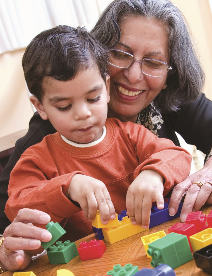 Abuela con gafas/lentes sentada con niño pequeño, ambos jugando con bloques lego/duplo de colores amarillo, rojo, y verde