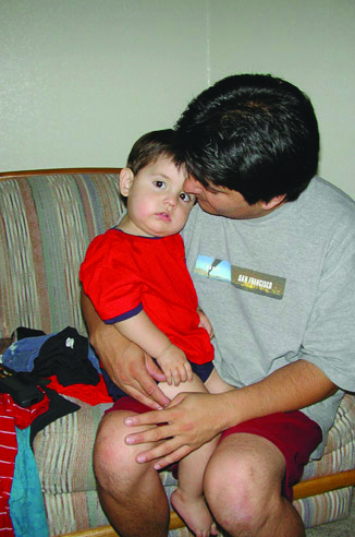 Papá sentado carga a su bebé y le sostiene su manita mientras el bebé apoya su cabeza en el hombro de su papá y observa sin sonreír