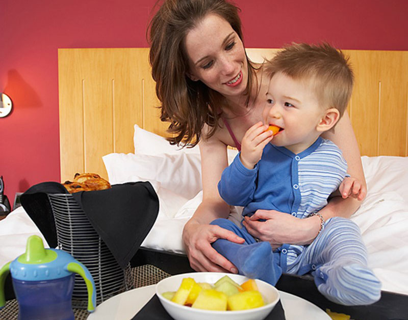 Un pequeño introduce un pedazo de fruta en su boca mientras su mamá lo carga sentados frente a una mesa con un plato de frutas