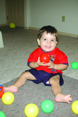 Niño pequeño sonriente, sentado sobre una alfombra rodeado de pequeñas pelotas plásticas de colores y sosteniendo con sus dos manos una pelota roja