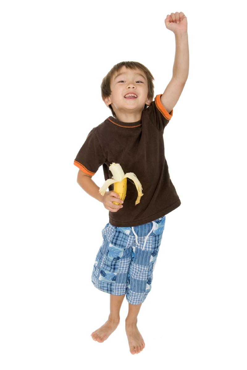 Niño pequeño sonriente sostiene y come una banana/plátano/cambur con una mano y levanta su otra mano