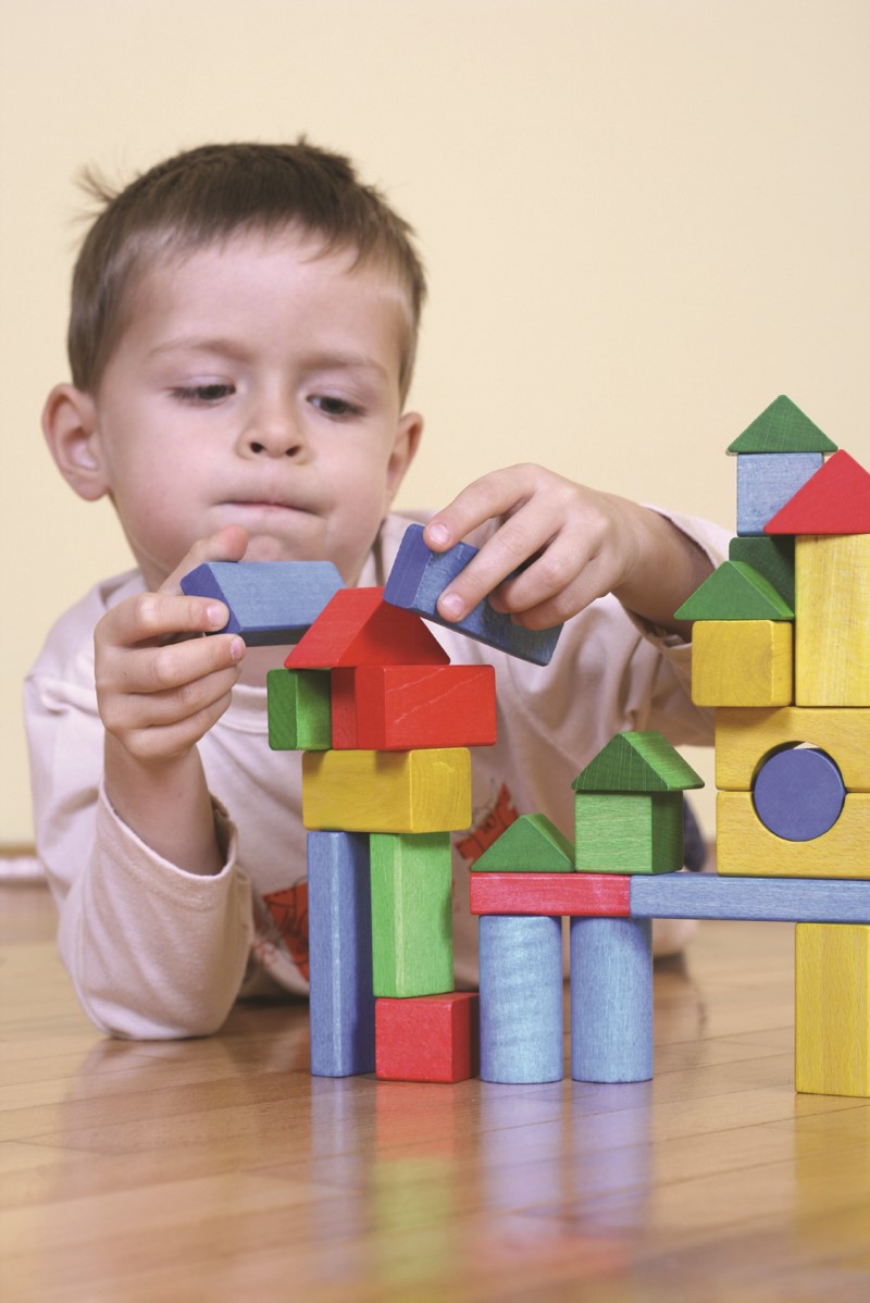 Niño pequeño sostiene dos piezas en sus manos mientras observa y construye una estructura con bloques de colores de madera