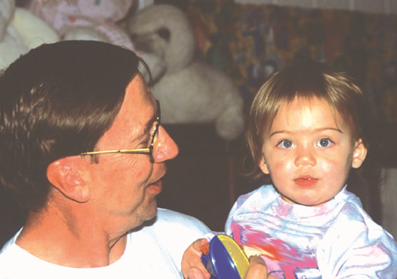 Papá con lentes/gafas mira con afecto a su niño a quien carga en sus brazos mientras él/ella sostiene un juguete sus manos