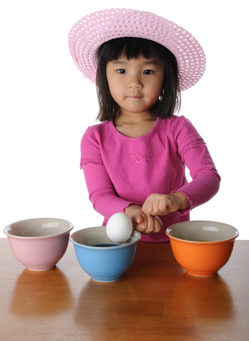 Niña pequeña con sombrero de ala sentada frente a tres bóles/recipientes de colores con  un huevo de Pascua