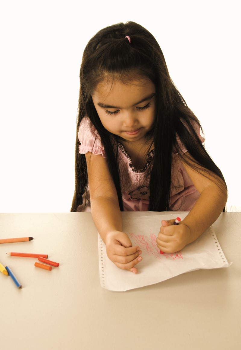 Niña pequeña traza líneas usando un creyón o crayola color rosa con una mano y sostiene el papel con la otra mano