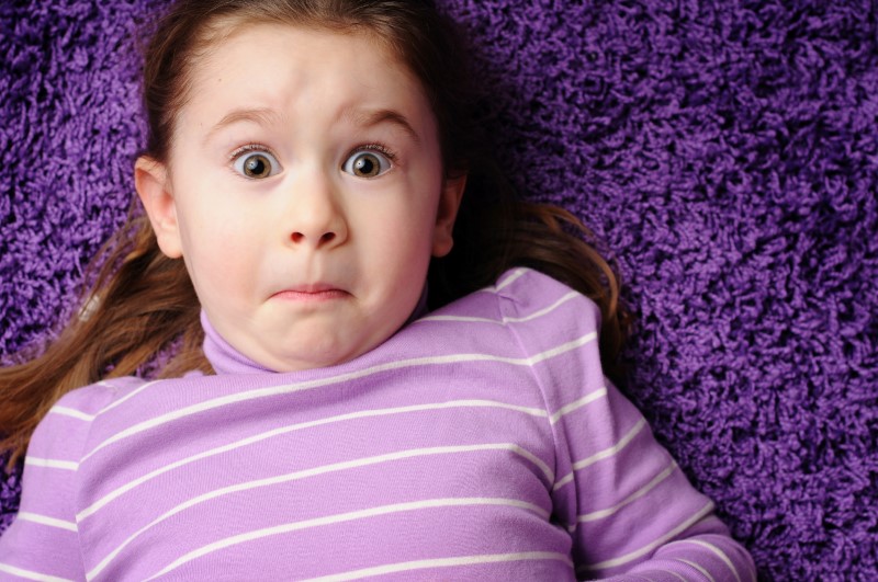 Niña pequeña acostada sobre una alfombra color violeta/morada mira fijamente con ojos abiertos y expresión de sorpresa