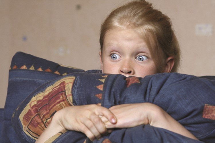 Niña pequeña con ojos muy abiertos detrás de un almohadón azul