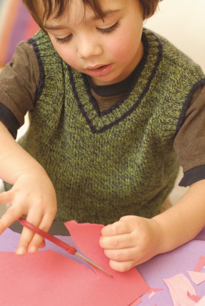 Niño pequeño recorta con una tijera la esquina de una hoja de cartulina/papel de construcción