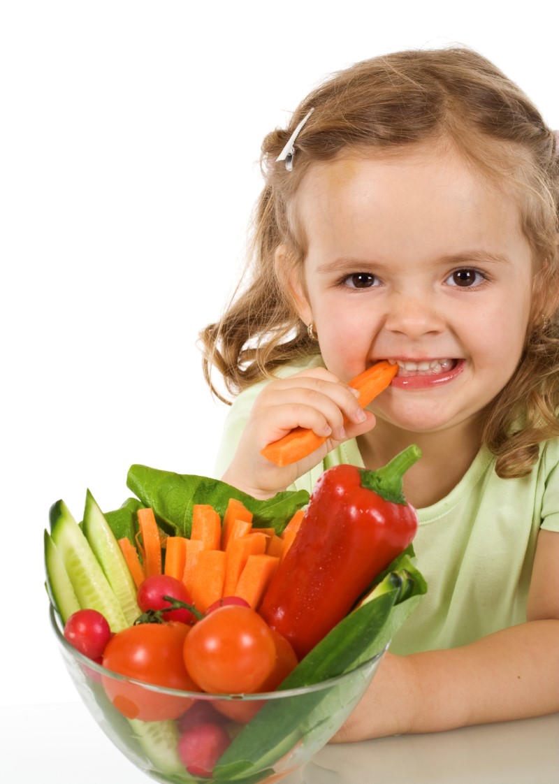 Niña pequeña frente a un bol con pepinos, pimenón, zanahorias y tomates listos para comer mientras muerde un trozo de zanahoria