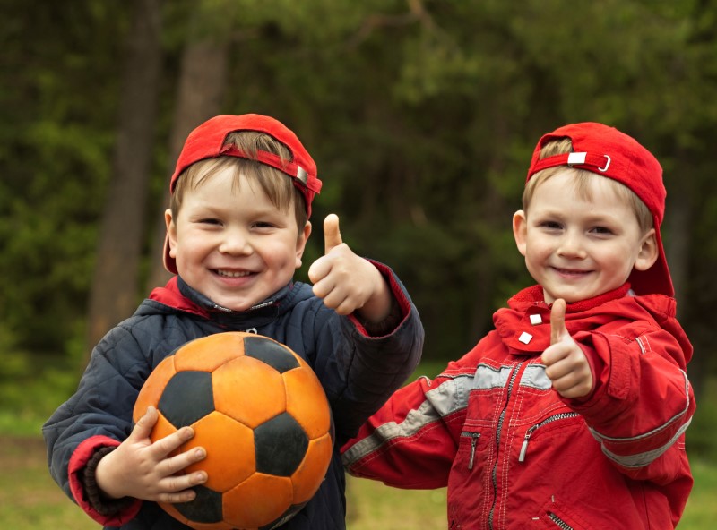 Dos niños pequeños sonríen y se abrazan en un campo deportivo mostrando sus pulgares en señal de aprobación mientras uno de ellos sostiene un balón de fútbol
