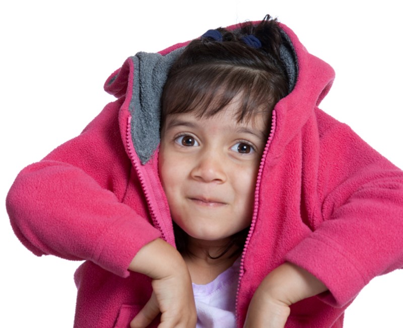 Niña pequeña cubre su cabeza mientras dobla sus brazos dentro de una chaqueta lanuda o fleecy color fuchsia  observando con mirada y sonrisa pícaras