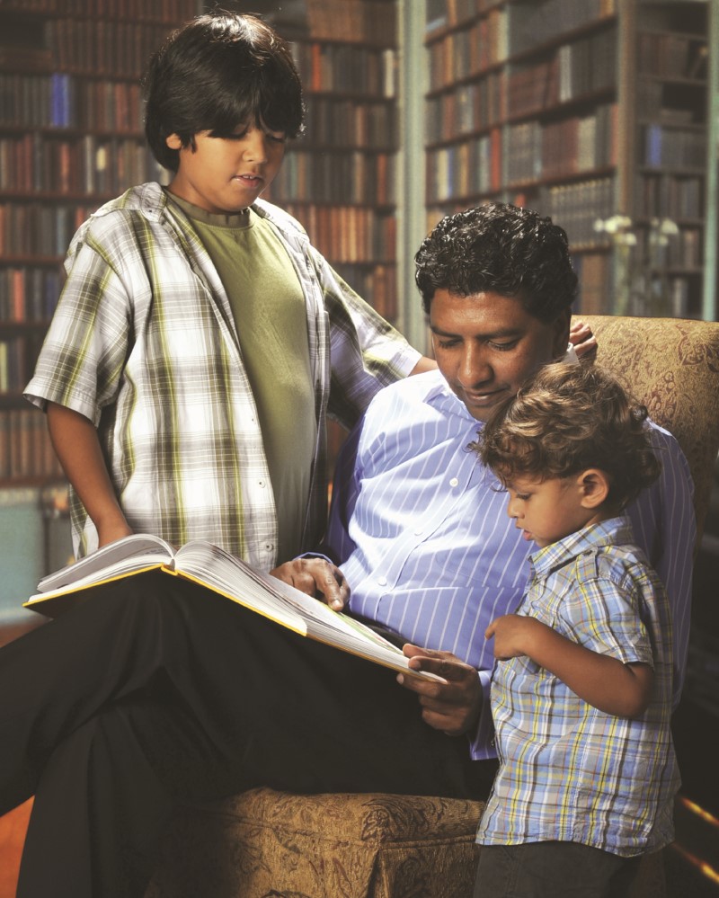 Papá sentado con dos niños edades entre 3 y 9 a 10 leyendo un libro en una biblioteca