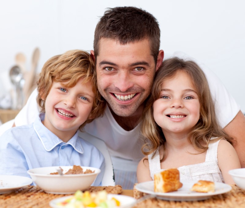 Papá abraza a su niño y a su niña en mientras toman su cereal, frutas y pastelitos