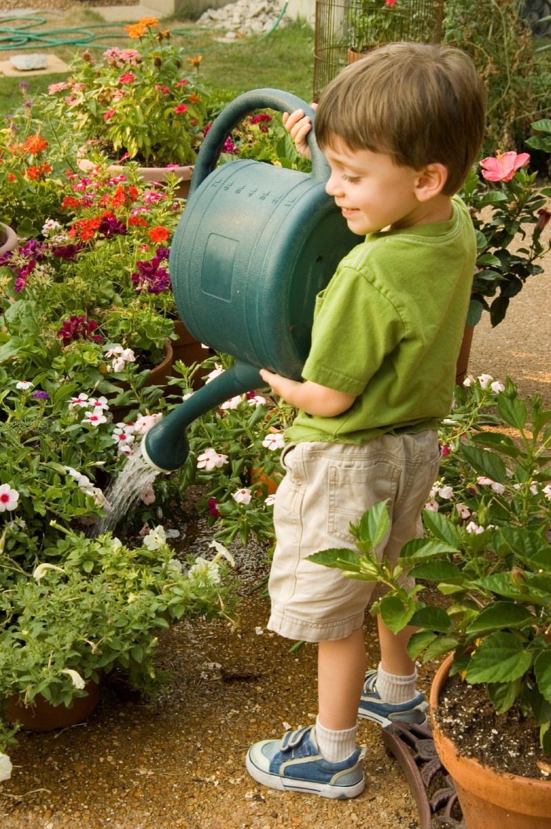 Niño pequeño sostiene una regadera verde en sus manos mientras riega en un patio las plantas con flores en sus macetas