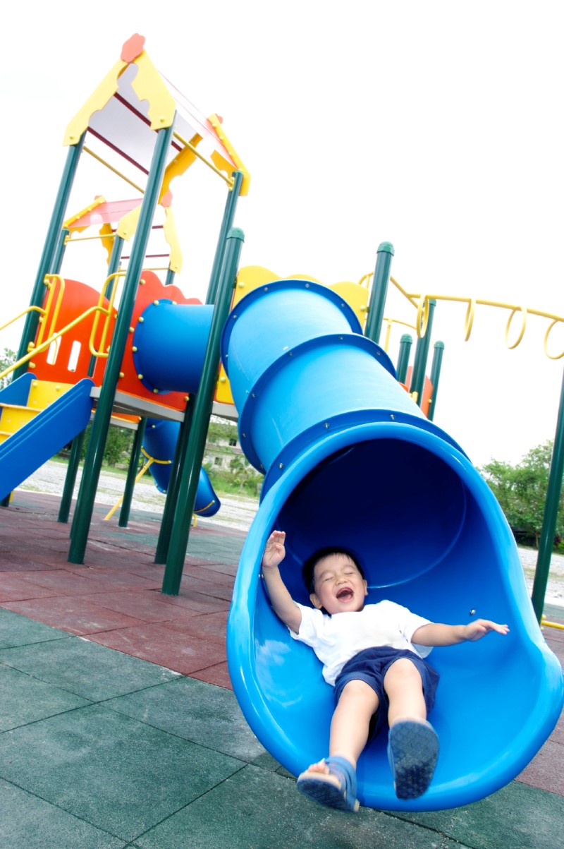 Niño pequeño sonríe mientras se desliza hacia el final de un tobogán/resbaladilla en un parque de juego
