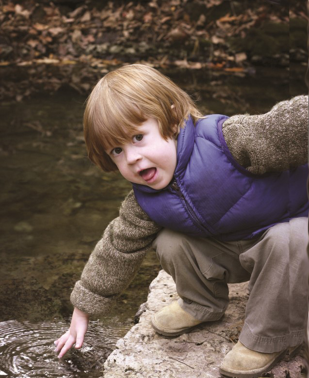 Niño pequeño agachado sobre una roca toca el agua de un río/lago mientras abre sus ojos y su boca con sorpresa